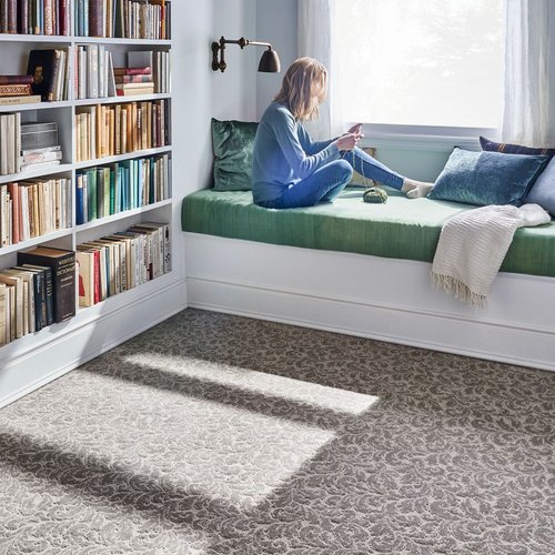 A woman knitting on a bed having a library next to her in a bedroom with brown carpet floor provided by Howard Carpenter Floor Covering in Danville KY